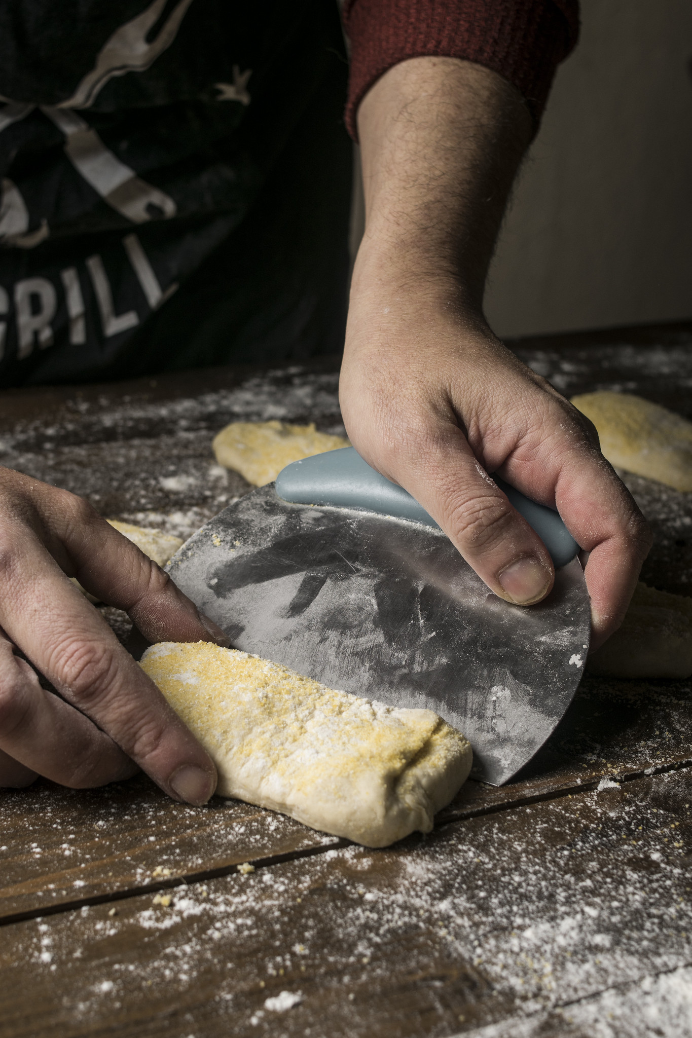 Hands using a dough scraper to divide wet ciabatta dough into rustic rolls on a flour-dusted wooden surface.